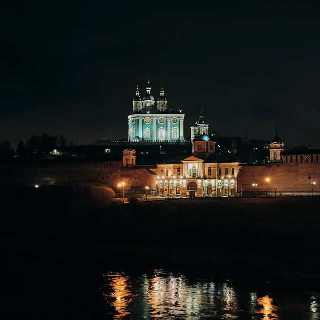 An illuminated cathedral above old walls, with lights reflected in the river.