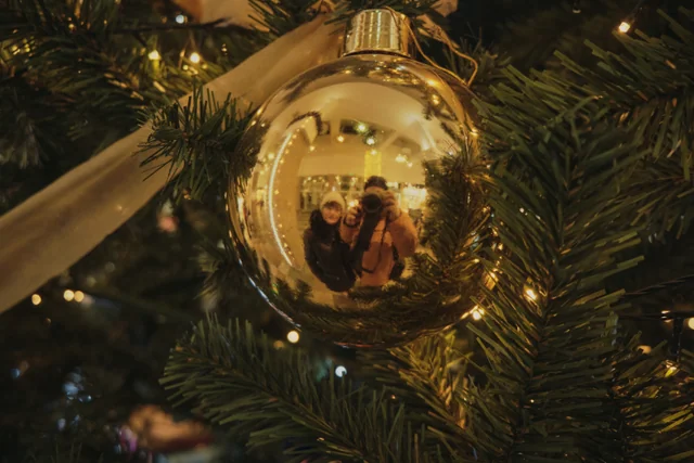 Two people, one holding a camera, reflected in a golden Christmas ornament.