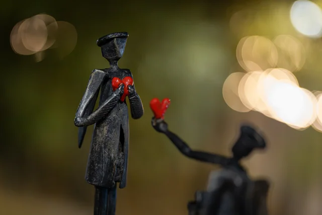 Two metal figures holding red hearts against softly blurred evening lights.