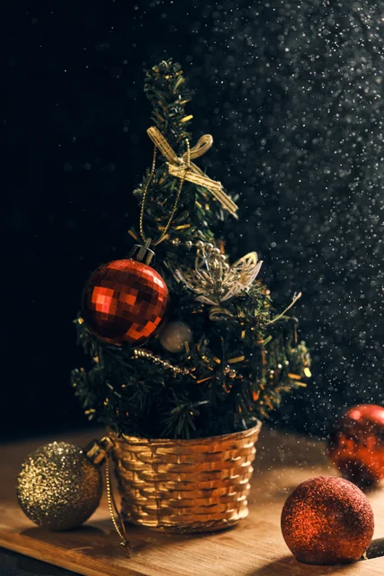 A small decorated Christmas tree in a woven basket beside red ornaments.