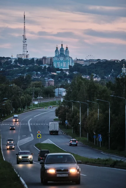 Cars follow a winding city road beneath a distant cathedral at sunset.