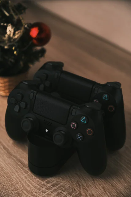 Two black game controllers on a wooden table beside a small decorated Christmas tree.